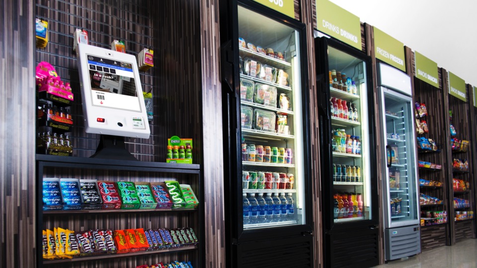 Die Selbstbedienungskasse Gen3 von 365 Retail Markets in einem Regal umgeben von Kassenvorsatz-/Impulsartikeln. Rechts anschließend stehen mehrere PicoCooler-Kühlschränke, gefühlt mit verschiedenen Produkten, sowie Regale mit weiteren Artikeln.